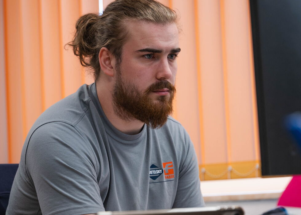 Ein bärtiger Mann mit Dutt arbeitet konzentriert am Laptop in einem Büro mit orangefarbenen Jalousien.