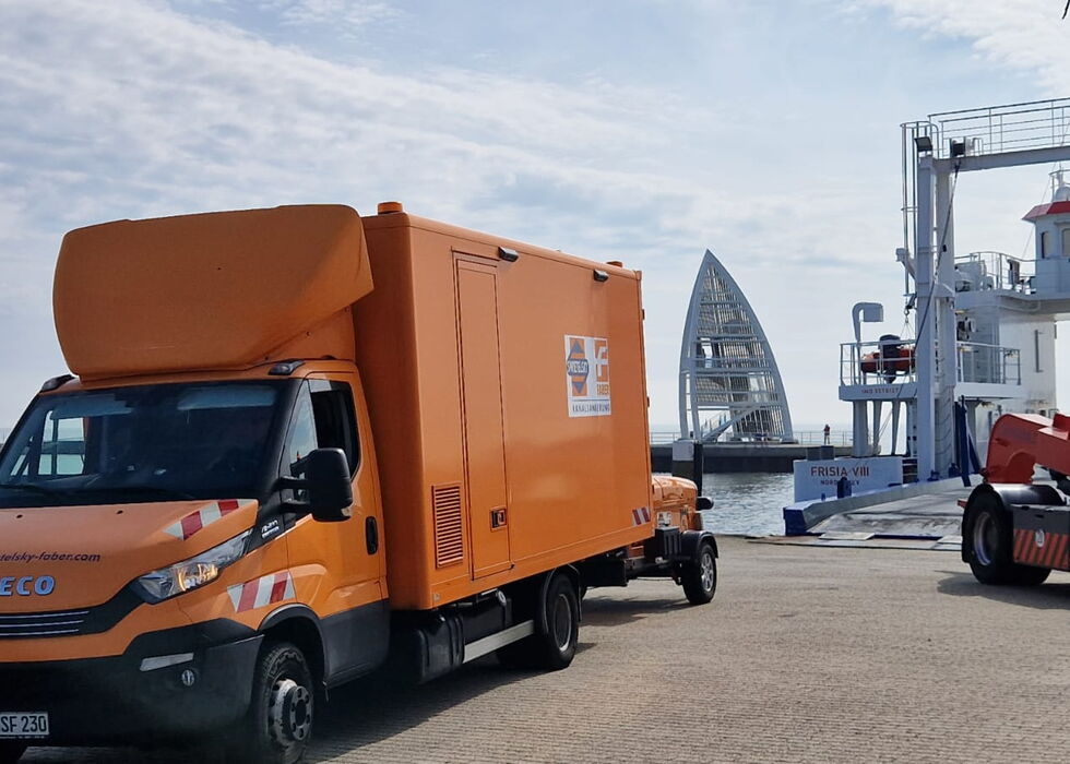 Oranger Servicelaster am Hafen geparkt, mit Kränen und einem segelförmigen Gebäude im Hintergrund.