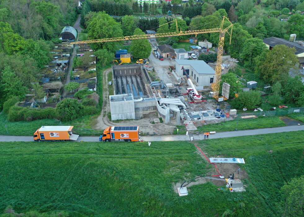Luftaufnahme einer Baustelle mit Kran, im Bau befindlichen Gebäuden und Lastwagen neben einer Straße, umgeben von Feldern und Bäumen.