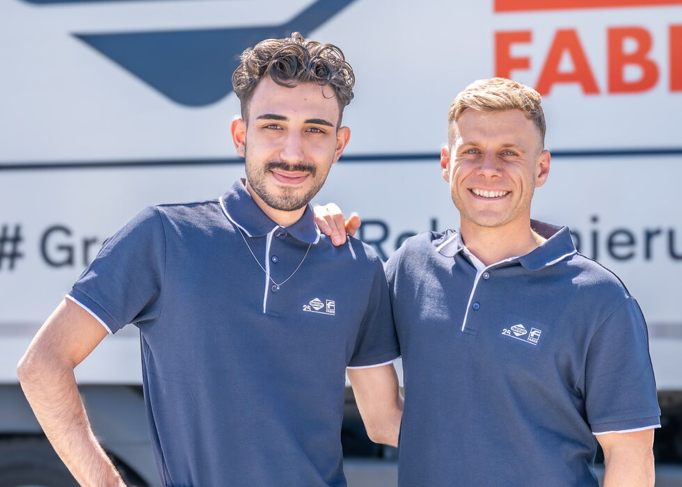 Zwei lächelnde Männer stehen in dunkelblauen Poloshirts vor einem beschrifteten Firmenwagen.