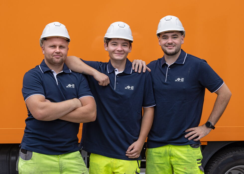 Drei Bauarbeiter mit Schutzhelmen und Arbeitshemden stehen vor einem orangefarbenen Lkw und lächeln in die Kamera.