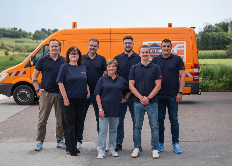 Sieben Personen stehen im Freien vor einem orangefarbenen Servicewagen.