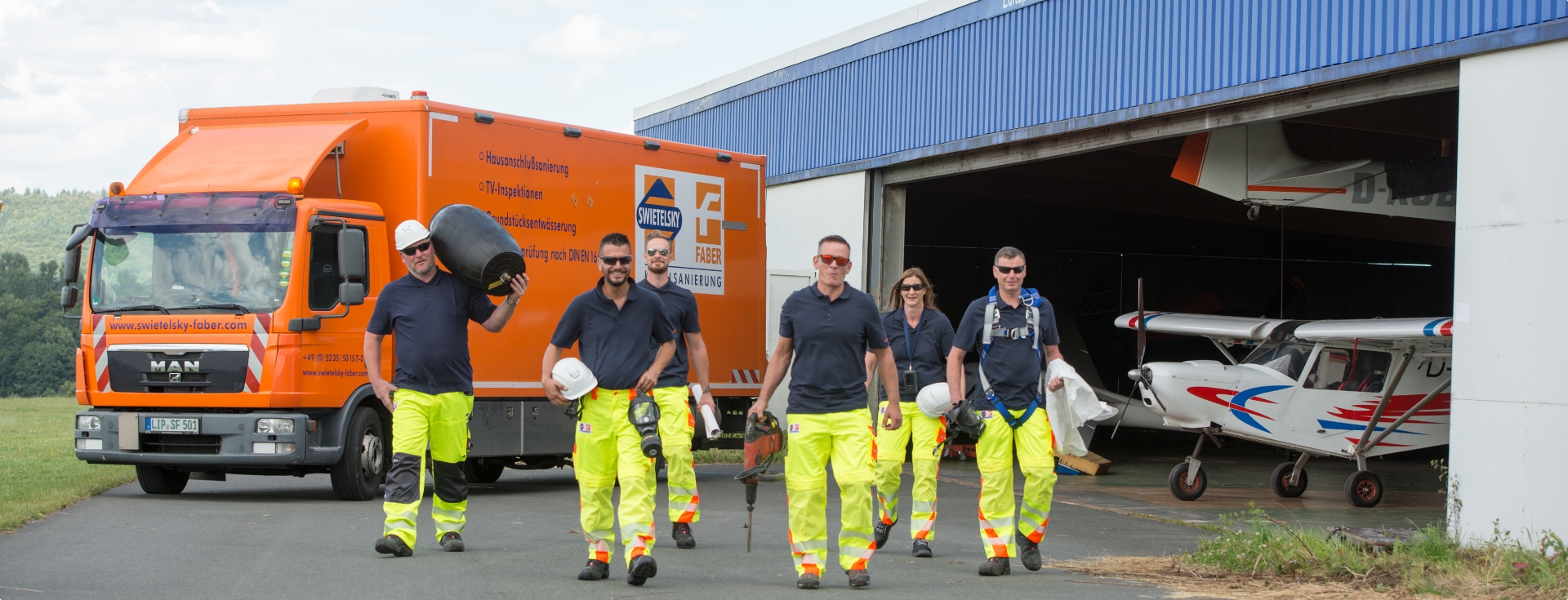 Eine Gruppe von fünf Arbeitern in Sicherheitskleidung geht auf die Kamera zu, mit einem orangefarbenen Lkw und Flugzeugen im Hintergrund.